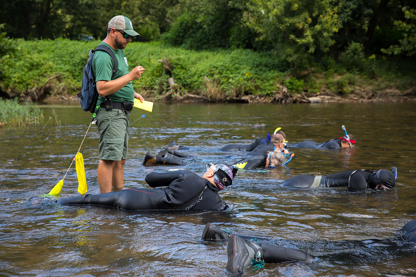 A group of people dressed in wet suits with snorkels on lie face-down in a shallow river while another person stands over them, recording data on a clipboard.