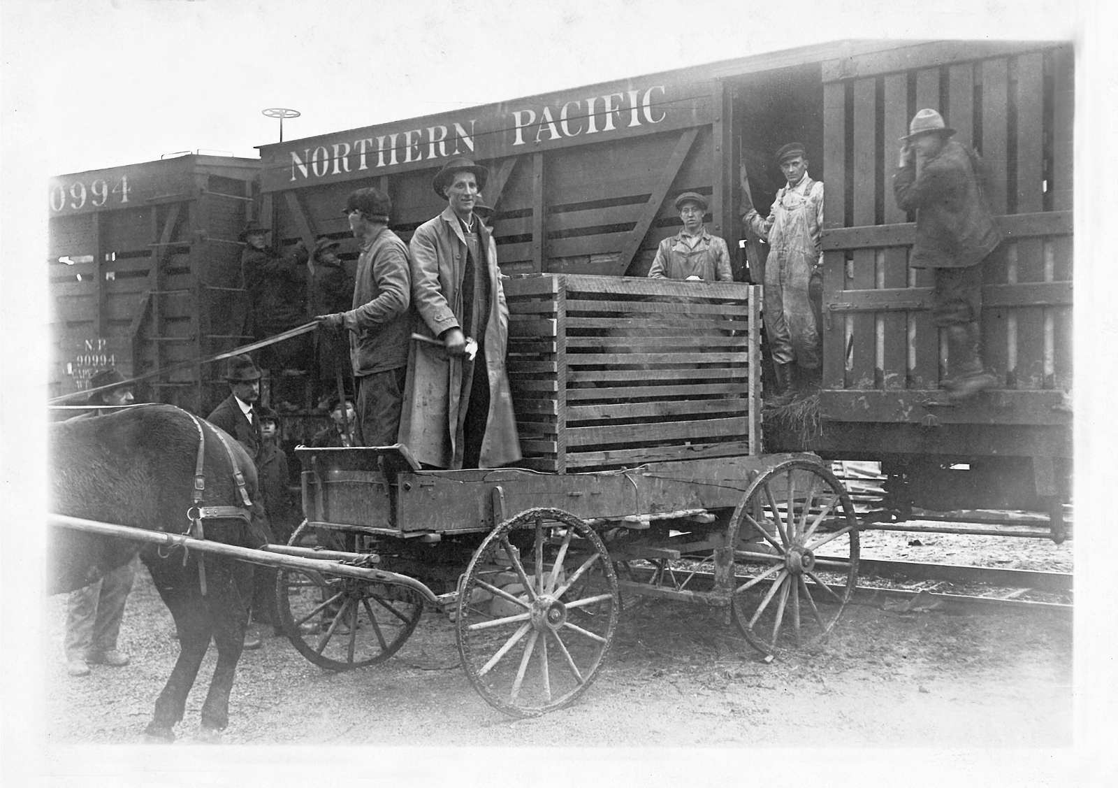 A historical black-and-white photo of four men on a horse-drawn wagon with a large crate backed up to a railroad car.