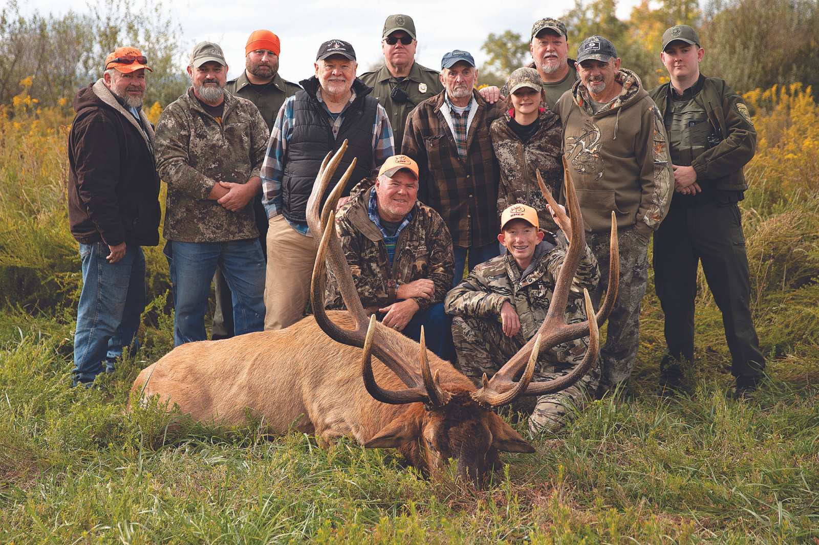 A large harvested bull elk lying on the groud, surrounded by a group of people.