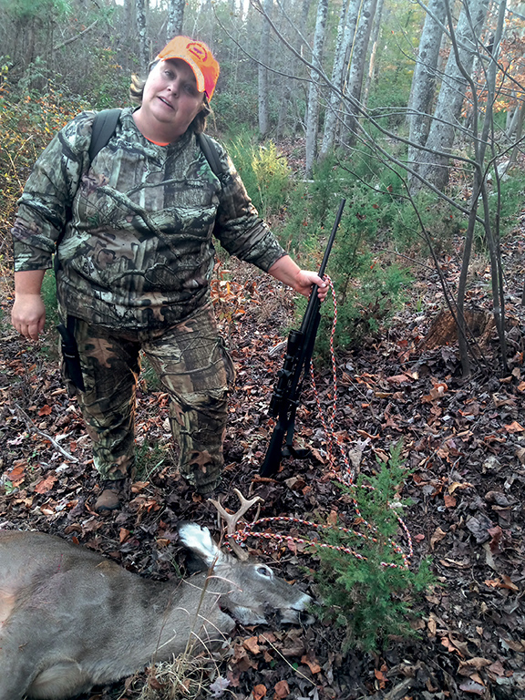 A photo of a woman in camouflage with a gun standing over a harvested buck.