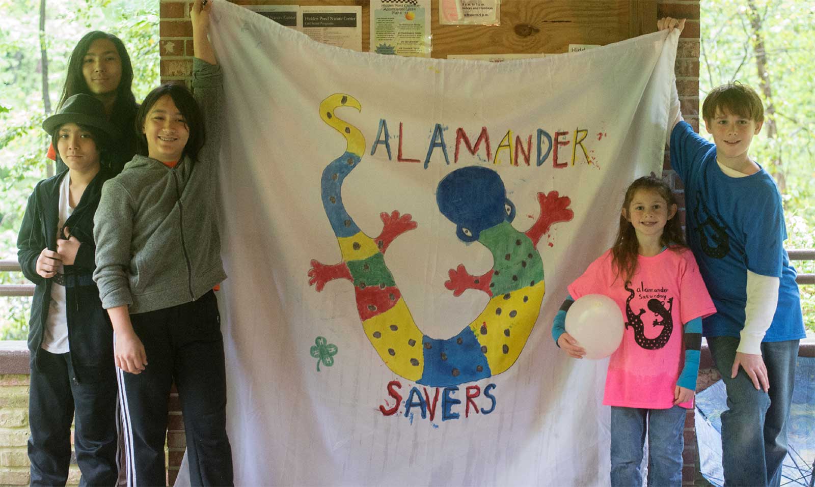A picture of several children holding up a sheet that says "salamander savers!" this is part of the youth conservation initiative to raise awareness about salamanders and their plights