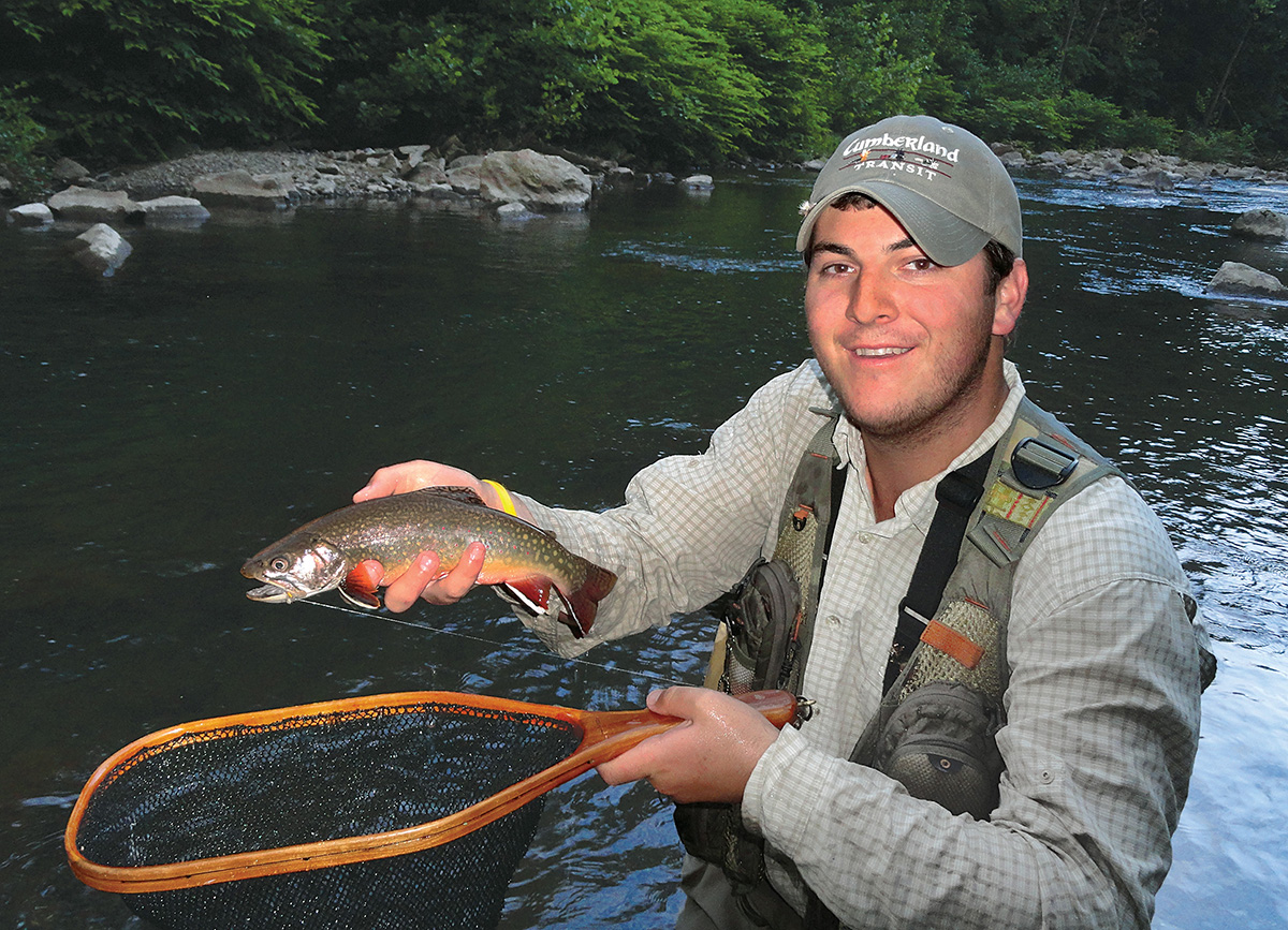 An image of a angler holding a net and a brown trout in front of a river