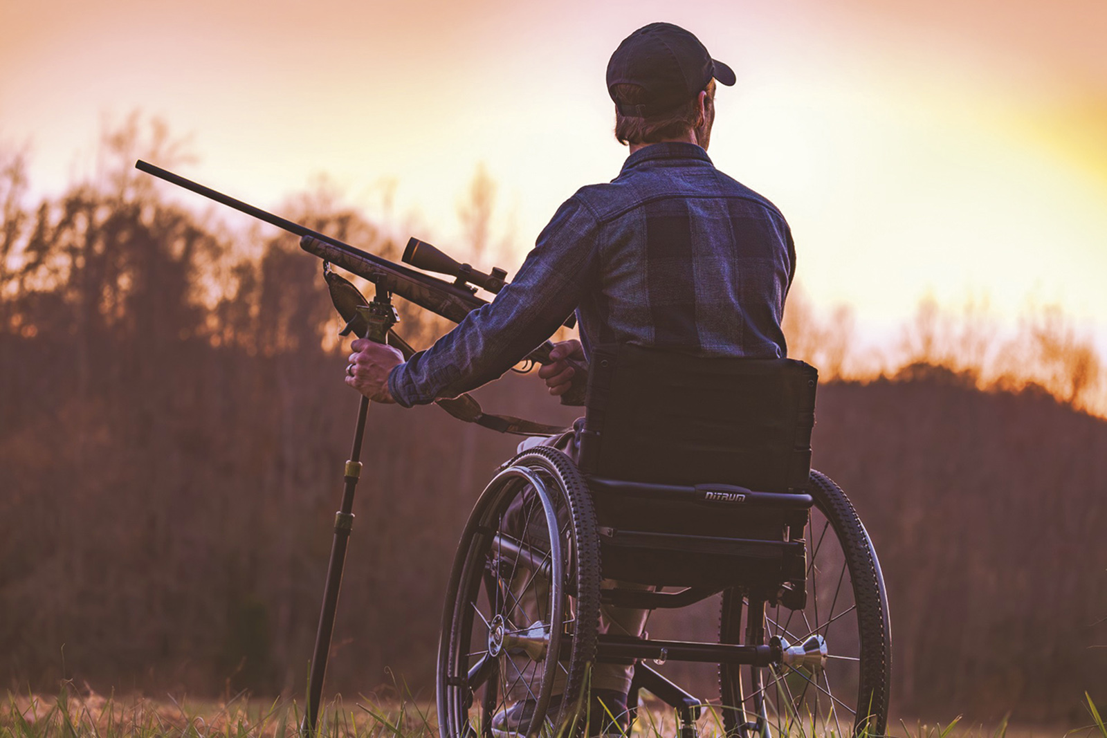 A photo taken at dusk with a hunter in a wheelchair holding a rifle silhouetted against a treeline.