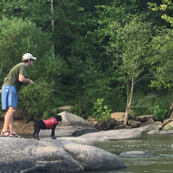 An image of a man fishing with a black lab wearing a red life vest named Waffles on a boulder in the James river
