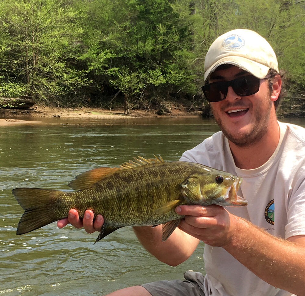 An image of a man with a white hat who is the Aquatic Education Coordinator for the DGIF holding a smallmouth bass with a river visible behind him.