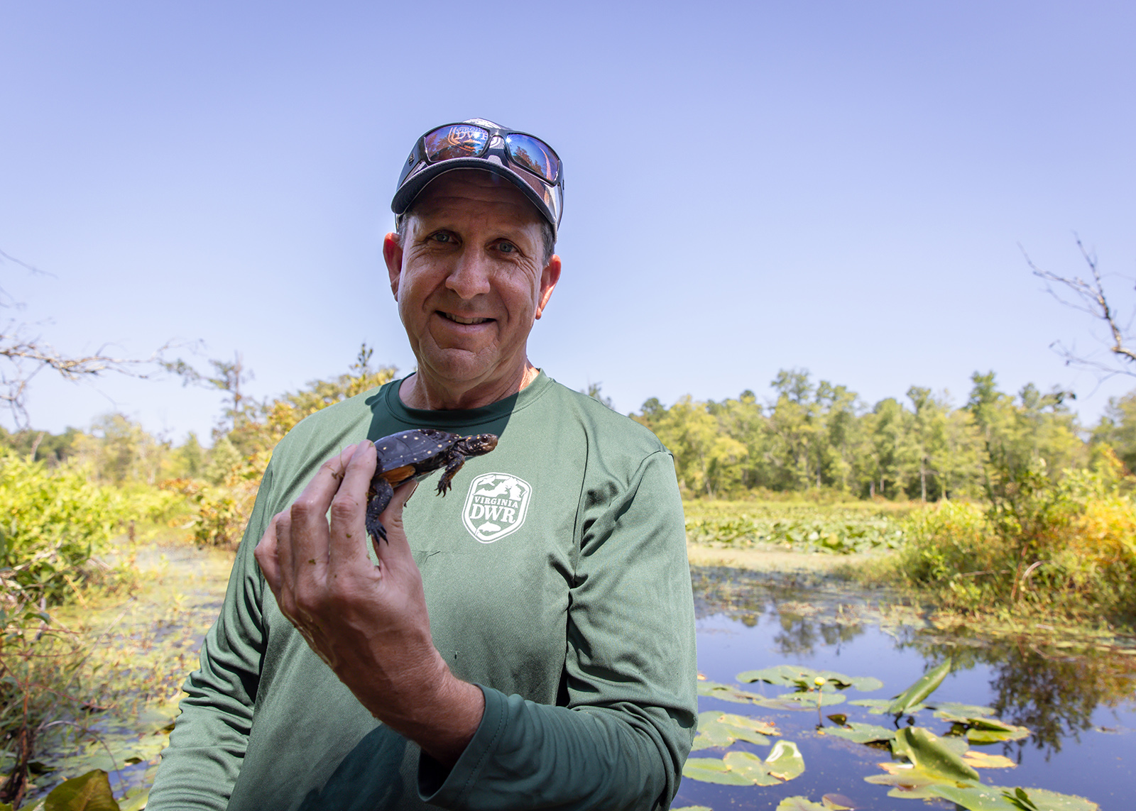 A photo of a man standing in front of a body of water in a DWR uniform shirt and holding a small spotted turtle in his hand.