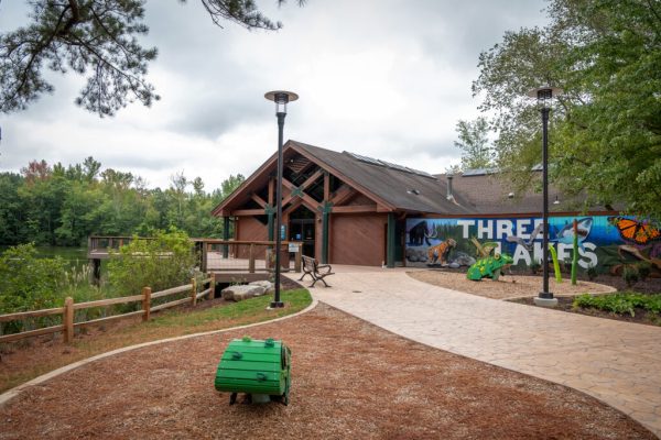 The front entrance to the Nature Center, which is a brown building that has a mural painted on one side that reads "Three Lakes." To the left of the building is a wooden observation deck that overlooks one of the lakes.