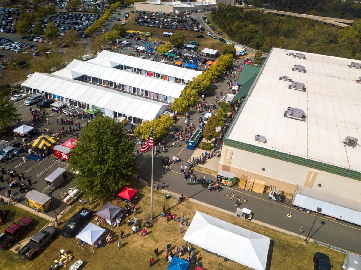 AN image of an event venue with multiple tents set up and a parking lot filled with cars in the background; in the foreground many people are present