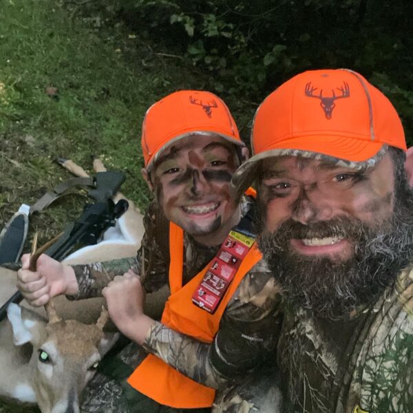 An man and a deer smiling at the camera with camouflage face paint and a deer the boy had killed