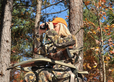 An image of a woman in a tree viewing platform with a rifle and binoculars looking to hunt deer